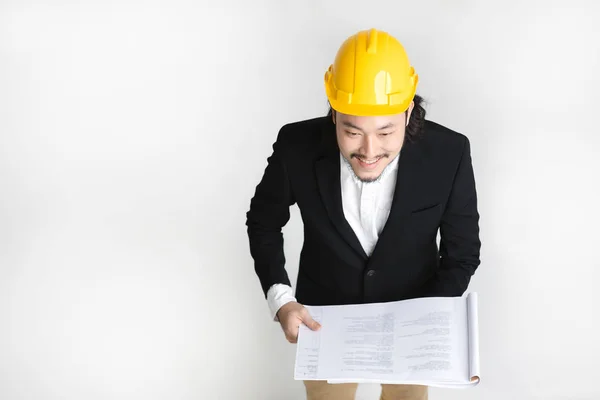 Top view of happy, young Asian business man in black suit and white shirt, wearing yellow helmet, looking upwards to camera, cheerily thumb-up, hold project plan in hand, on white isolated background