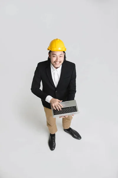 黒いスーツと白いシャツを着た幸せな若いアジアのビジネスマンのトップビュー 黄色のヘルメットを着用し カメラを上向きに見て 手にノートパソコンのキーボードで入力し 白い孤立した背景に — ストック写真