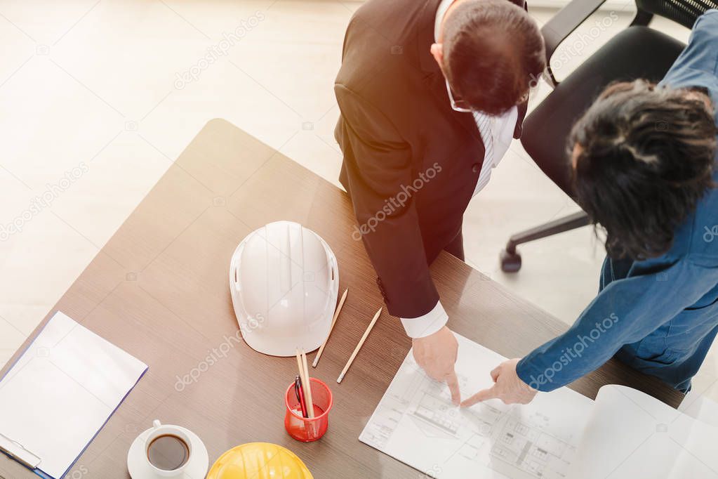 Close up, top view of Asian business man and male foreman in coverall, discussing at drawing, building plan design, with laptop, helmet on working desk.