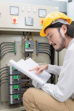 Genç Asyalı elektrik mühendisi bej pantolon ve beyaz gömlek, yakın çekim, Sarı kask, öğretim el çalışma Denetim Masası bodur takmak, onun rutin günlük kontrol izleyin