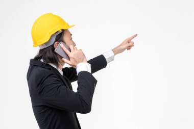 Asyalı mühendis portresi, siyah takım elbiseli, başında sarı kask, ayakta ve duvara işaret, cep telefonu, önünde beyaz duvar kullanın.