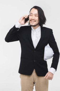 Genç Asyalı mühendis portresi , siyah takım elbiseli, cep telefonu kullanın, kolunda beyaz kask, ayakta, arkasında beyaz arka plan.