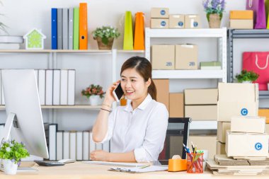 Güzel, çekici Asyalı iş kadını at kuyruklu, beyaz gömlekli, bilgisayarın önünde oturan, gülümseyen yüzlü, akıllı telefonu tutan, ofisin girişinde arayan.