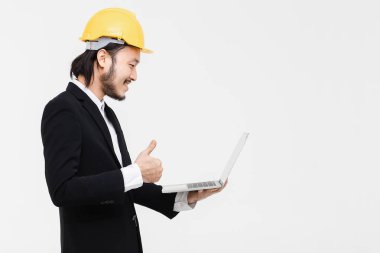 Asyalı mühendisportresi, siyah takım elbiseli, başında sarı kask, ayakta, kolunda defter tutun ve onu kullanarak, arkasında beyaz arka plan.
