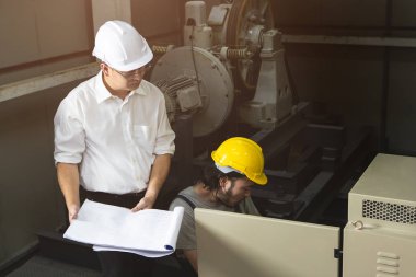 Orta yaş Asya fabrika yöneticisi Görüntü, elinde eğitim çalışma, kask giyen tulum genç Asyalı mühendis konuşurken, kontrol paneli önünde çömelmek, ağır makine arka plan ile