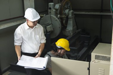 Orta yaş Asya fabrika yöneticisi Görüntü, elinde eğitim çalışma, kask giyen tulum genç Asyalı mühendis konuşurken, kontrol paneli önünde çömelmek, ağır makine arka plan ile