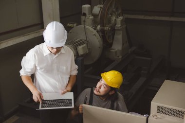 Orta yaş Asya Fabrika Müdürü, el, bir dizüstü bilgisayar ve miğfer, Denetim Masası önünde bodur tulum Genç Asyalı mühendisi ağır makine arka plan sorunları tartışmak