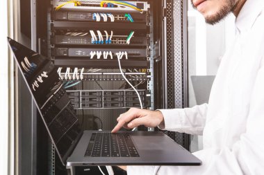 Bej pantolon ve beyaz gömlek li mühendisyakın, kontrol paneli önünde oturan, elinde dizüstü bilgisayar da yazarak, onun rutin günlük kontrol takip