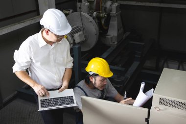 Orta yaş Asya Fabrika Müdürü, el, bir dizüstü bilgisayar ve miğfer, Denetim Masası önünde bodur tulum Genç Asyalı mühendisi ağır makine arka plan sorunları tartışmak