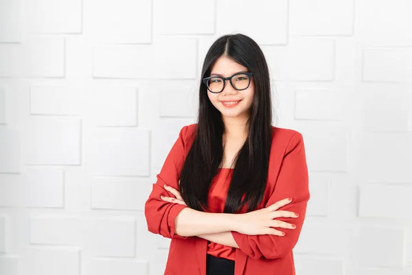Portrait of young attractive Asian businesswoman, wearing glasses, long black hair in red suit and black pant, crossed arms, smile with satisfaction, look at camera on white brick wall background