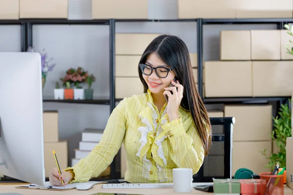 Young attractive Asian businesswoman, long black hair, wearing glasses, yellow blouse, talking on smartphone with customer, taking note, look at computer in startup office background full of boxes