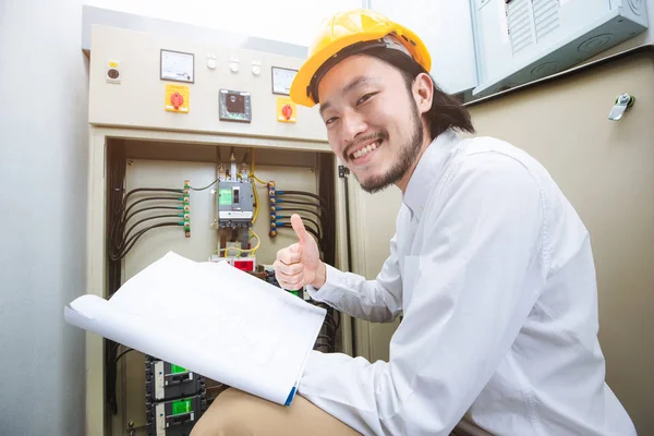 Close up of young Asian electric engineer in beige pant and white shirt, wear yellow helmet, squat near control panel, working instruction in hand, follow up his routine daily checking