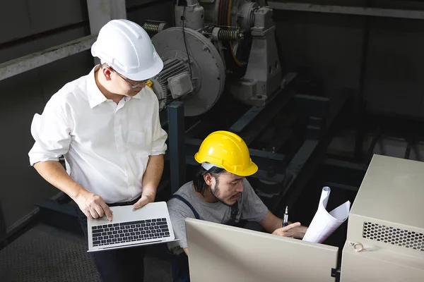 Middle age Asian factory manager, laptop in hand, and young Asian engineer in jumpsuit wearing helmet, squat in front of control panel, discuss problems with heavy machinery background