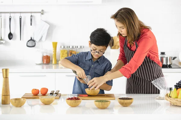 Asian mom was teaching her son, pouring spice powder in a wooden container, intently, in a clean, white and modern kitchen.