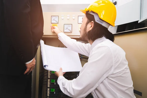 Young Asian engineer in beige pant and white shirt, wear yellow helmet, squat near control panel, explain, report to middle age manager in business suit, standing next to him