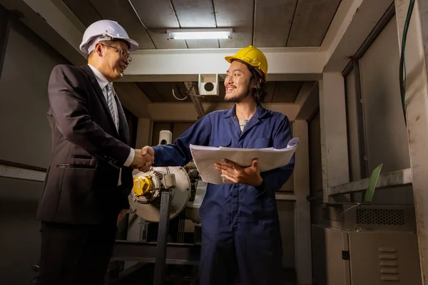 Young Asian engineer handshakes with executive engineer beside heavy machinery in industrial factory, blueprint in his hand, concept for boss comes visit industrial factory.