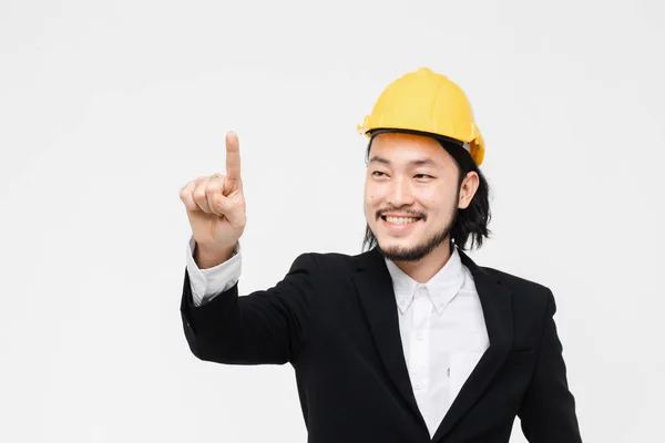Portrait of Asian engineer, in black suit, yellow helmet on his head, standing and point come forward, white background behind him.