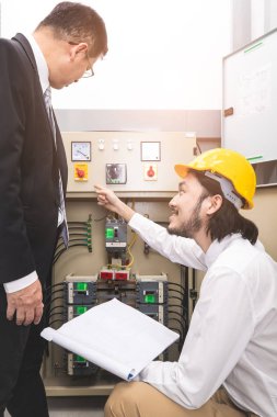 Bej pantolonlu ve beyaz gömlekli genç Asyalı mühendis, sarı kask takıyor, kontrol panelinin yanında çömeliyor, açıkla, iş elbisesi içinde orta yaşlı yöneticiye, yanında duran