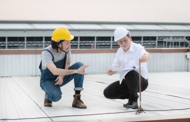 Orta yaşlı Asyalı fabrika müdürü ve kot tulumlu genç Asyalı mühendis, güvenlik ayakkabıları, miğferler, üretim binasının çatısında Şimşek Çubuğu durumu, dizüstü bilgisayar.