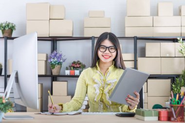 Young attractive Asian businesswoman, long black hair, wearing glasses, yellow blouse, recheck order  and message from customer, taking note, in startup office background full of boxes clipart