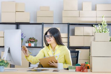Young attractive Asian businesswoman, long black hair, wearing glasses, yellow blouse, recheck order  and message from customer, taking note, in startup office background full of boxes clipart