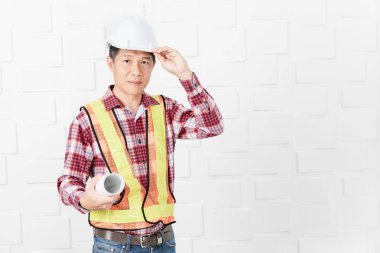 Turuncu, sarı yansıtıcı Emanet yelek, kol, beyaz tuğla duvar ofiste ayakta altında holding beyaz kask giymiş orta yaşlı Asya yakışıklı mimar, kot pantolon ve ekose desenli gömlek,