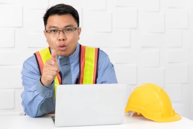 Shot Orta yaşlı Asya, tulum site mühendisi, turuncu giyen, sarı yansıtıcı güvenlik yeleği, çene altında el, proje hakkında düşünme, dizüstü bilgisayar ile, beyaz ofis arka plan sarı güvenlik şapkası