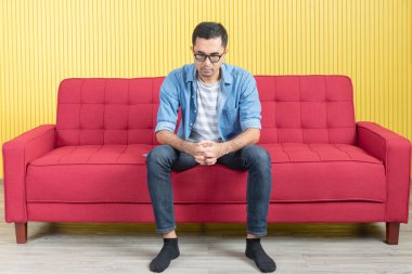 Genç Asyalı yakışıklı sakallı adam close-up, kucaklayışını oturan kırmızı koltuk, ciddi bir yüz, eller üzerinde kot gömlek giyen gözlük chin, modern oturma odasında altında parlak sarı çizgili duvar arka plan