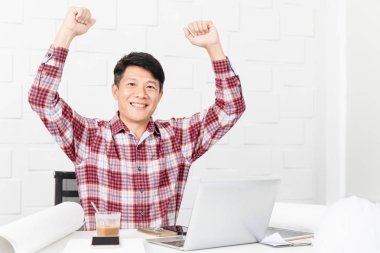 Yumruk kadar dizüstü bilgisayar, kask, tablet, kahve ve belgeleri arka planda tutan kazanan jest yapma inşaat site ofiste çalışan Asya yakışıklı mimar, ekose desenli gömlek, orta yaşlı