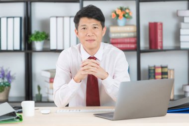 Orta yaşlı, yakışıklı, Asya, işadamı, beyaz gömlek, koyu kırmızı kravat, başlangıç ofisinde oturan eller, çalışma masası, dizüstü bilgisayar ve kahve Kupası arka plan üzerinde gururla poz geçti