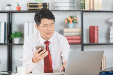 Başlangıç ofisinde çalışan, akıllı telefon tutarak, müşteri adı listesi denetimi yaşında, yakışıklı, Asya, işadamı, koyu kırmızı kravat, Beyaz gömlekli orta ve satınalma siparişi defter üzerinde