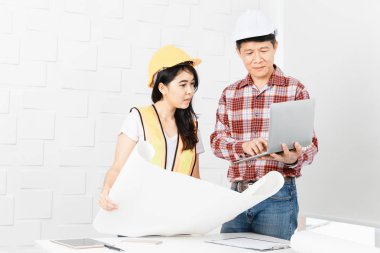 Veri üstünde laptop genç kadın Asya Yardımcısı gösterilen, portakal, sarı yansıtıcı Emanet yelek ve kask, planları elinde giyen orta yaşlı Asya mimar, kot pantolon ve ekose desenli gömlek,