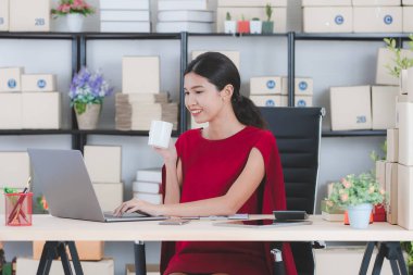 İş istasyonunda oturan yüz, gülümseyen genç güzel, çekici Asya işkadını Kırmızı elbiseli portresi e-postalar, modern başlangıç office arka plan denetimi sırasında kahve sabah fincan keyfini çıkarın