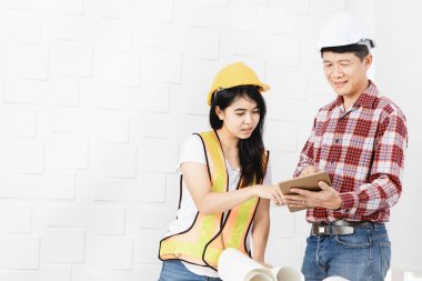 Orta yaşlı Asyalı mimar, kot pantolon ve kontrol gömlek, genç kadın Asyalı asistan tablet verileri açıklamak, turuncu giyiyor, sarı yansıtıcı güvenlik yeleği, ve sarı hardhat, ofis arka plan