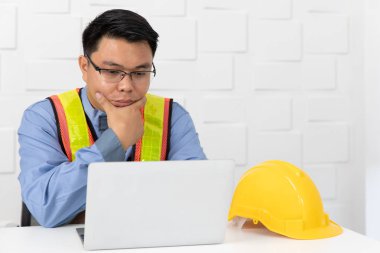 Shot Orta yaşlı Asya, tulum site mühendisi, turuncu giyen, sarı yansıtıcı güvenlik yeleği, çene altında el, proje hakkında düşünme, dizüstü bilgisayar ile, beyaz ofis arka plan sarı güvenlik şapkası