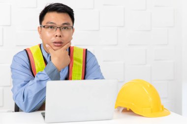 Shot Orta yaşlı Asya, tulum site mühendisi, turuncu giyen, sarı yansıtıcı güvenlik yeleği, çene altında el, proje hakkında düşünme, dizüstü bilgisayar ile, beyaz ofis arka plan sarı güvenlik şapkası