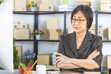 Asyalı, üst düzey iş kadını, kısa saçlı, gözlük takan, iş istasyonunda oturan, kolunu çapraz, ev ofis arka plan ile oder bilgisayara bakıyor Shot