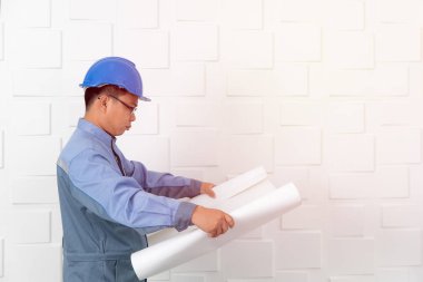 Orta yaşlı, Asya elektrikçi veya içinde tulum, üniforma, giyiyor mavi emniyet kask, çalışma İnşaat çizim, taslakları, beyaz ofis arka planda çalışan teknisyeni varoluş portresi