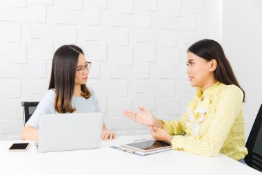 Two young, beautiful, Asian architects, on informal meeting, discussing about project management timelines, using tablet and notebook, at construction site boardroom, with white brick wall background clipart