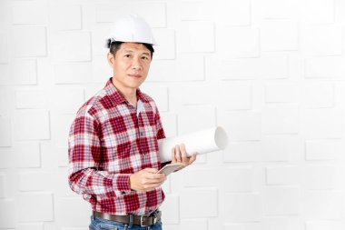 Giydiği beyaz kask, ayakta, kullanım smartphone, orta yaşlı Asya yakışıklı mimar, kot pantolon ve ekose desenli gömlek, beyaz duvar arka plan üzerinde kol altında inşaat planları rulo