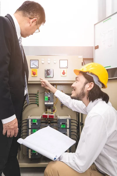 Bej pantolonlu ve beyaz gömlekli genç Asyalı mühendis, sarı kask takıyor, kontrol panelinin yanında çömeliyor, açıkla, iş elbisesi içinde orta yaşlı yöneticiye, yanında duran