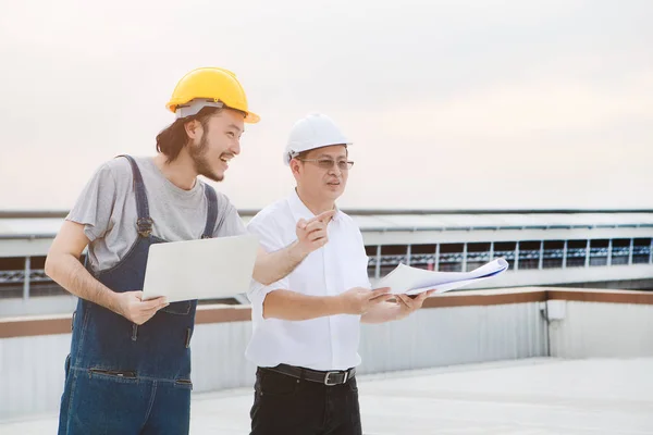 Middle age Asian factory manager and young Asian engineer in jeans jumpsuit, safety shoes, helmets, checking Lightning Rod condition on the rooftop of manufacturing building, laptop in hand