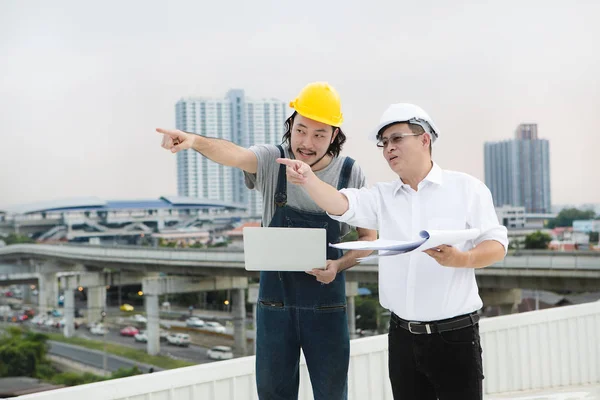 Middle age Asian factory manager and young Asian engineer in jeans jumpsuit, safety shoes, helmets, checking Lightning Rod condition on the rooftop of manufacturing building, laptop in hand