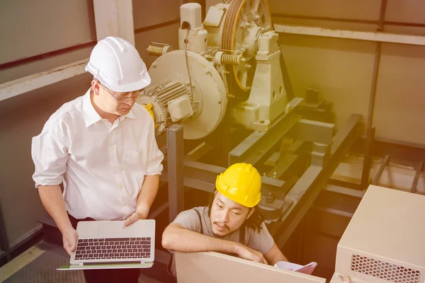 Orta yaş Asya Fabrika Müdürü, el, bir dizüstü bilgisayar ve miğfer, Denetim Masası önünde bodur tulum Genç Asyalı mühendisi ağır makine arka plan sorunları tartışmak