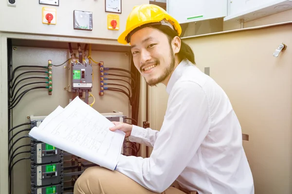 Close up of young Asian electric engineer in beige pant and white shirt, wear yellow helmet, squat near control panel, working instruction in hand, follow up his routine daily checking
