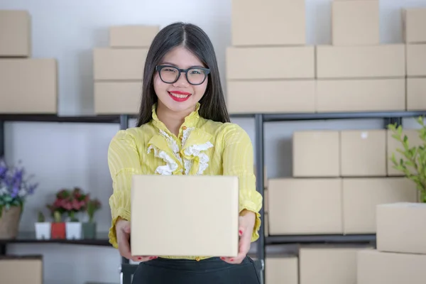 Young attractive Asian businesswoman, long black hair, wearing glasses, yellow blouse, smiling face, show package ready to send off to customer, in startup office background full of boxes
