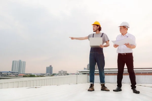 Middle age Asian factory manager and young Asian engineer in jeans jumpsuit, safety shoes, helmet, point something on far away on the rooftop of manufacturing plant