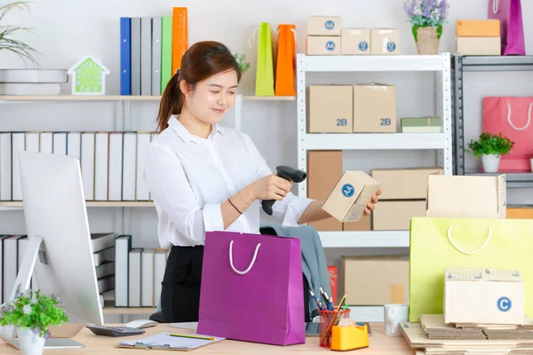 Genç, çekici Asyalı iş kadını, beyaz gömlek, gülümseyen yüz, pakette barkod okuyucu kullan. Mor alışveriş çantası ve başlangıç ofisi geçmişi olan müşteriye göndermeye hazır.