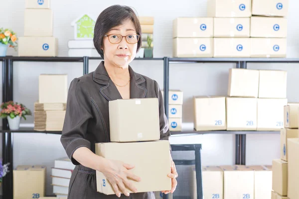 Portrait of Asian, senior businesswoman, short hair, wearing eyeglasses, casual suit, standing in home office, holding packages ready to send off to customers, look at camera