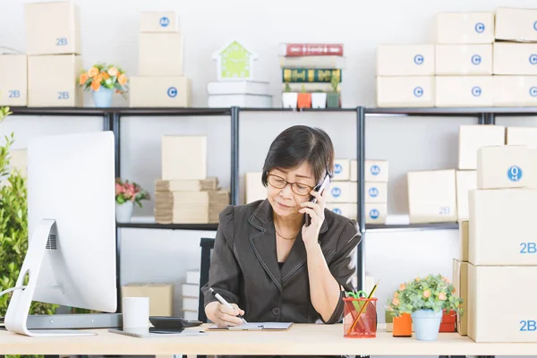 Studio portrait of Asian, senior businesswoman, short hair, wearing eyeglasses, sitting and writing at her working desk, pointing at computer screen, with home office background